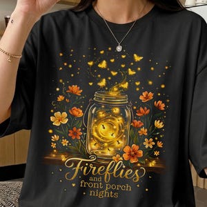Może przedstawiać: Czarny t-shirt z motywem słoika wypełnionego świecącymi świetlikami, otoczonego pomarańczowymi i żółtymi kwiatami i motylami. Tekst "Fireflies and front porch nights" jest napisany ozdobną czcionką.
