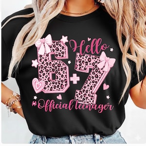 May include: Black t-shirt with a pink leopard print design featuring the numbers 67, a plus sign, and the words "Hello" and "Official teenager." The design includes pink bows, hearts, and stars.