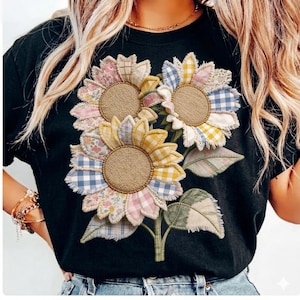Puede incluir: Camiseta negra con una aplicación de tres girasoles. Los girasoles tienen centros de arpillera y pétalos de tela de retazos con estampados florales, cuadros y lisos. Hojas y tallos verdes completan el diseño.