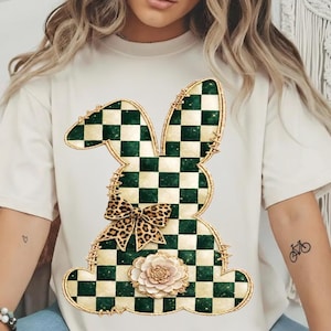 May include: Cream-colored t-shirt featuring a large bunny design. The bunny is a patchwork of green and cream checkered patterns, outlined in gold. A leopard print bow and a cream-colored flower are also part of the design.