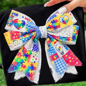 May include: A large, colorful bow featuring a patchwork design with puzzle pieces, hearts, and polka dots. The bow has a white lace trim and a blue center. The bow is held against a black background.