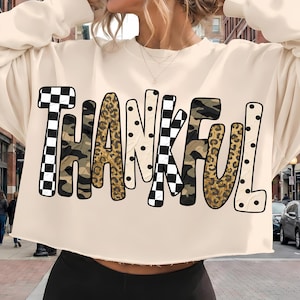 May include: A cream-colored long-sleeve sweatshirt with a raw-edge hem. The front features the word THANKFUL in large, stylized letters, each filled with a different pattern including black and white checkers, leopard print, camo, and polka dots.