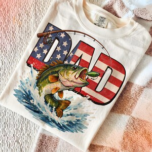 Black shirt mockup with a bold patriotic fishing dad PNG design showing a largemouth bass biting a lure over American flag DAD lettering, great for outdoor dad and fisherman apparel