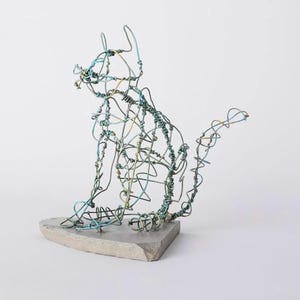 May include: A wire sculpture of a cat in a seated position. The cat is made of thin, intertwined metal wires in shades of blue and green, mounted on a grey, stone-like base. The sculpture has a minimalist, artistic aesthetic.