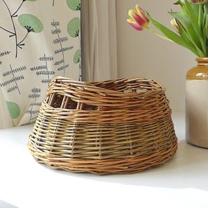 May include: A handcrafted willow basket with a flared design, showcasing natural tones. The basket sits on a white surface, with a ceramic vase holding tulips in the background. The basket's dimensions are approximately 25 cm in diameter.