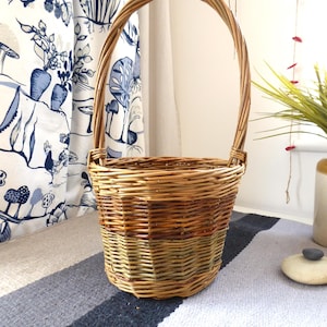 May include: A hand-woven wicker basket with a tall handle. The basket features a mix of light and dark brown tones, with a band of green near the base. The handle arches gracefully above the basket's opening. The basket is on a striped rug.