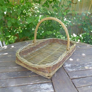 May include: A rectangular wicker basket with a handle. The basket is made of woven light brown and tan willow branches. The handle arches over the top. The basket is on a weathered wooden table.