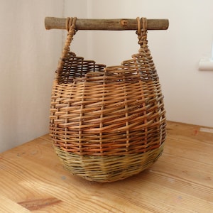 May include: A woven basket featuring a rounded body made of light brown and green willow branches. The basket has an open top and a horizontal handle constructed from a single, straight, natural wooden branch secured with woven willow strips.