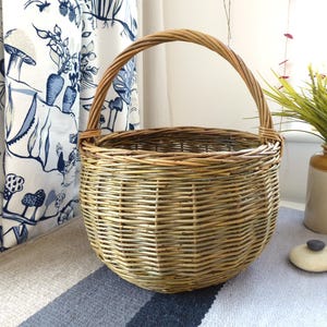 May include: A round, woven wicker basket with a curved handle. The basket is light brown and tan. It is made of woven wicker. The basket is on a striped surface, with a curtain in the background.