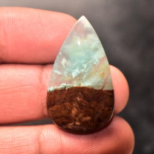 Puede incluir: Una gema en forma de lágrima con un patrón de color único. La mitad superior presenta una mezcla de azul claro, blanco y toques de amarillo, mientras que la mitad inferior es de un marrón oscuro intenso. La piedra se sostiene en una mano.