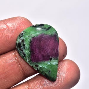 Könnte beinhalten: Ein polierter Edelstein in Tropfenform. Der Stein hat einen zentralen, tiefmagentafarbenen Bereich, der von einer leuchtend grünen Matrix mit schwarzen Sprenkeln umgeben ist. Der Edelstein wird in einer Hand gehalten.