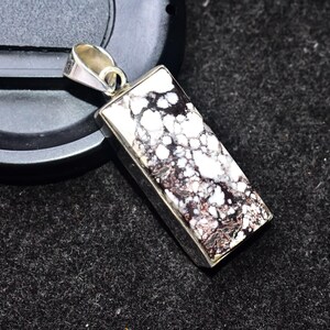 Peut inclure: Pendentif rectangulaire avec un cadre argenté. Le pendentif présente une pierre tachetée blanche et marron foncé. La bélière est également argentée. Le pendentif est présenté sur une surface gris foncé.