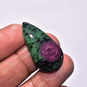 Könnte beinhalten: Ein polierter, tropfenförmiger Edelstein mit einer lebendigen Farbkombination. Der Stein zeigt eine tiefmagentafarbene Mitte, umgeben von einem gesprenkelten grün-schwarzen Muster. Der Edelstein wird in einer Hand gehalten, was seine glatte, reflektierende Oberfläche hervorhebt.