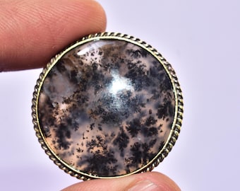 May include: A round cabochon gemstone featuring a translucent, milky white base with intricate, dark brown to black dendritic fern-like inclusions, set within a decorative, twisted rope-style metal border frame.
