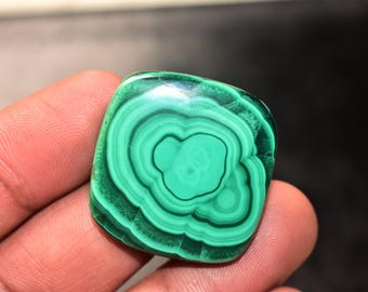 May include: A polished, square-shaped malachite gemstone. The stone displays concentric, swirling patterns of varying shades of green, creating a striking visual effect. The gemstone is held in a hand.