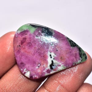 Könnte beinhalten: Ein polierter Edelstein mit einer Mischung aus Farben. Der Stein weist einen leuchtenden Magenton auf, mit Bereichen in Grün, Schwarz und Weiß. Die Oberfläche ist glatt und reflektierend und zeigt die natürlichen Muster und Texturen des Minerals.