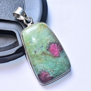 May include: A rectangular pendant with a silver-coloured bail. The pendant features a green stone with red inclusions, set in a silver bezel. The stone has a mottled appearance. The pendant is displayed against a white background.