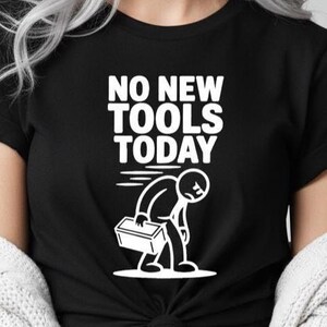 Könnte beinhalten: Schwarzes T-Shirt mit dem weißen Text "NO NEW TOOLS TODAY". Eine weiße Grafik zeigt eine niedergeschlagene Figur mit einer Werkzeugkiste. Das T-Shirt ist an der Taille geknotet.