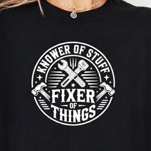 Op de afbeelding: Zwart T-shirt met een witte opdruk met de tekst "Knower of Stuff, Fixer of Things". Het ontwerp toont gekruiste sleutels en hamers in een cirkelvormig embleem. Het shirt wordt gedragen met een geruit overhemd.