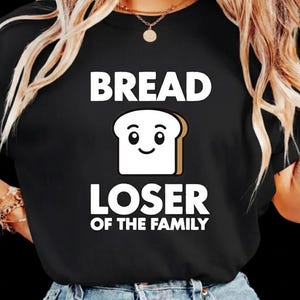 Op de afbeelding: Zwart t-shirt met de witte tekst "BREAD LOSER OF THE FAMILY". Het ontwerp bevat een lachende cartoon broodsnede. Een casual kledingstuk.