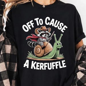 Peut inclure: T-shirt noir avec un motif fantaisiste. Un raton laveur portant un chapeau de cowboy et une cape chevauche un escargot, avec le texte "OFF TO CAUSE A KERFUFFLE" en blanc. Le design est sur fond noir.