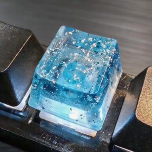 May include: Close-up of a mechanical keyboard with custom keycaps. One keycap is a translucent blue resin with silver flakes, and the others are black and metallic. The keyboard is on a dark surface.
