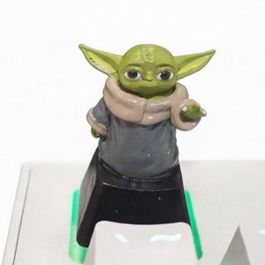 May include: A green and tan figurine of a character from a popular science fiction franchise. The figurine is standing on a white keyboard with green and blue accents. The character is wearing a gray robe and a tan scarf.