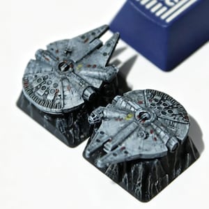 May include: Two gray Millennium Falcon keycaps with detailed weathering sit on black bases. A blue ESC keycap is in the background. The keycaps are designed for a mechanical keyboard, perfect for Star Wars fans.