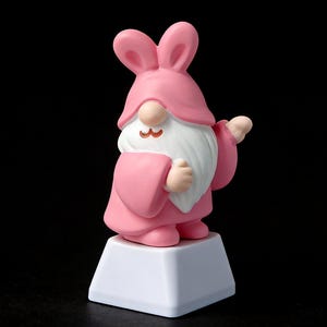 Può includere: Una statuina di gnomo rosa con un cappello a orecchie di coniglio e una veste, in piedi su una base quadrata bianca. Lo gnomo ha una barba bianca e piccoli baffi marroni. Lo sfondo è nero.