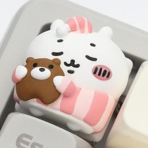 Puede incluir: Una tecla de teclado con un personaje de dibujos animados blanco y rosa con un oso de peluche marrón. La tecla está en un teclado de ordenador. El personaje lleva un gorro de dormir y un pijama a rayas. Diseño lindo y caprichoso.