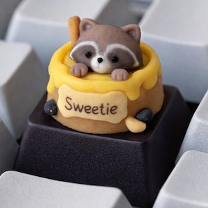 May include: A custom keycap featuring a raccoon in a honey pot design. The keycap is brown, yellow, and black, with the word "Sweetie" on a label. The keycap is set on a black keyboard key, surrounded by white keys.