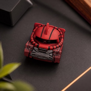 May include: A red and black artisan mechanical keyboard keycap shaped like a robotic head. The design features a central fin, a horizontal visor, and a textured dark gray chest plate, with black piping details encircling the rounded helmet.