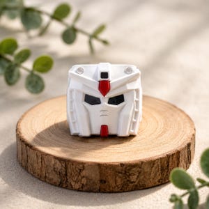 May include: A white robotic head figurine featuring a geometric design with angular panels, a central red crest on the forehead, a small red chin detail, and dark recessed eye sockets. The object has a symmetrical structure with two small circular accents.