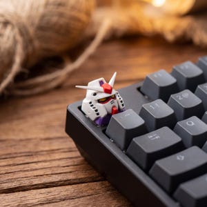 May include: A custom mechanical keyboard keycap shaped like a white robot head with red, yellow, and purple accents. The keycap is installed on a black keyboard, replacing the escape key, featuring a pointed crest and detailed mechanical vents.