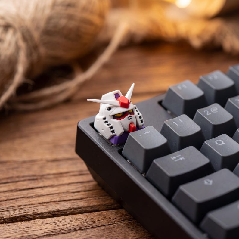 May include: A custom mechanical keyboard keycap shaped like a white robot head with red, yellow, and purple accents. The keycap is installed on a black keyboard, replacing the escape key, featuring a pointed crest and detailed mechanical vents.