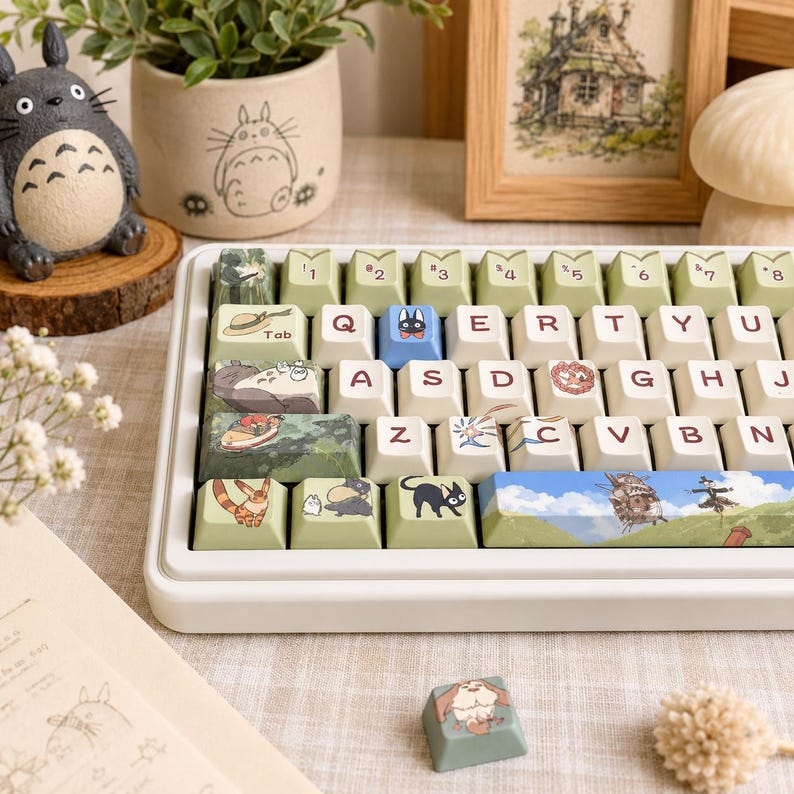 May include: A mechanical keyboard with custom keycaps featuring various animated character illustrations, including a black cat, a large grey creature, and a walking castle. The keys are a mix of cream, light green, and blue, with standard QWERTY layout.