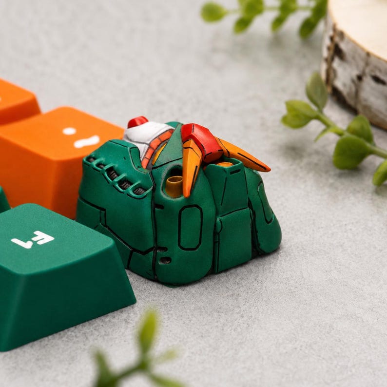 May include: A custom artisan mechanical keyboard keycap shaped like a green robotic torso with orange accents and a white collar. The keycap is positioned next to a solid orange keycap and a dark green keycap marked with the white text F1.