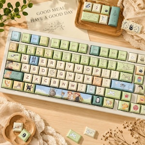 May include: A mechanical keyboard featuring light green and cream-colored keycaps decorated with various anime-style character illustrations and symbols. The spacebar displays a colorful scene with multiple figures, and additional loose keycaps are scattered nearby.