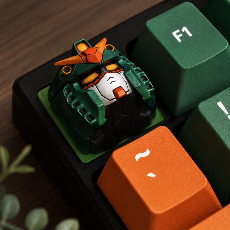 May include: A custom artisan mechanical keyboard keycap shaped like a stylized robot head in dark green, white, and orange. The keycap features a detailed faceplate with yellow eyes and a V-shaped crest, positioned between a green F1 key and an orange key.