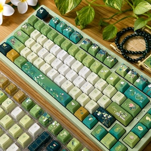 May include: A mechanical keyboard featuring a green frame and a set of custom keycaps in various shades of green, white, and dark teal. The keycaps display botanical illustrations, lotus flowers, and small insects, with standard QWERTY lettering.