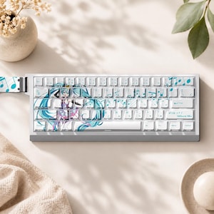 May include: A white mechanical keyboard featuring a graphic of the character Hatsune Miku with long teal hair, musical notes, and Japanese text. The keys display both English letters and Japanese characters, with a clean, light-coloured frame design.