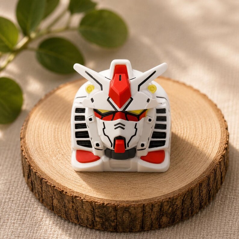 May include: A white, red, and yellow mechanical robot head keycap featuring a stylized face with black panel lines, a central red crest, and side vents. The keycap has a square base and a detailed, angular design inspired by mecha aesthetics.