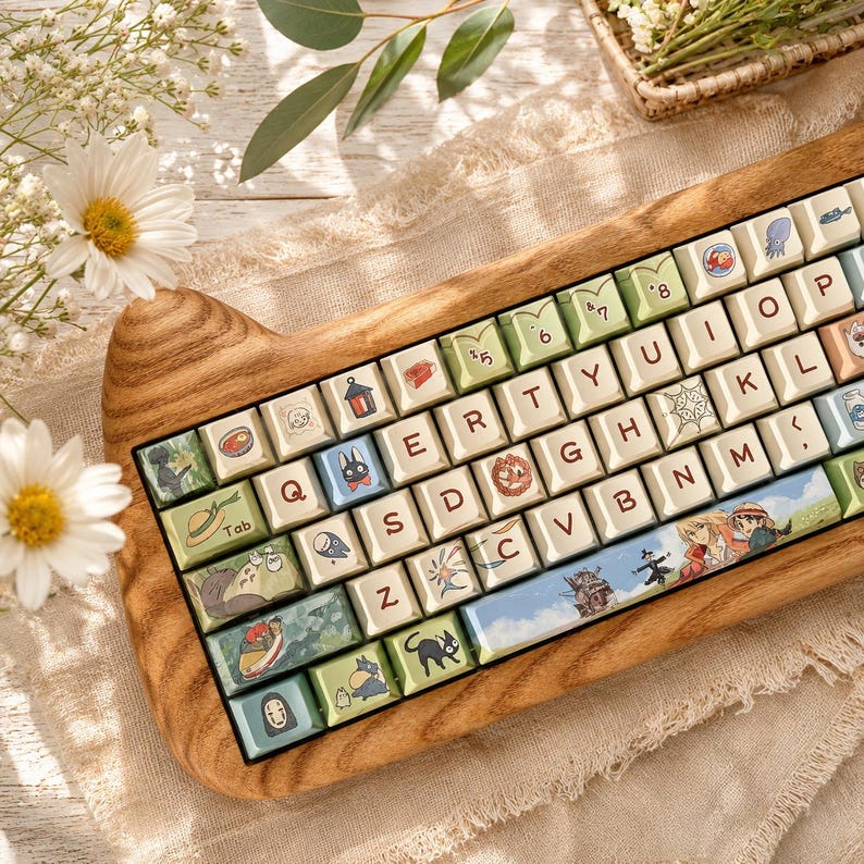 May include: A mechanical keyboard with a wooden case shaped like cat ears. The keycaps are cream-colored with dark brown letters and feature various colorful illustrations of animated characters, including a black cat and a castle, across the keys.