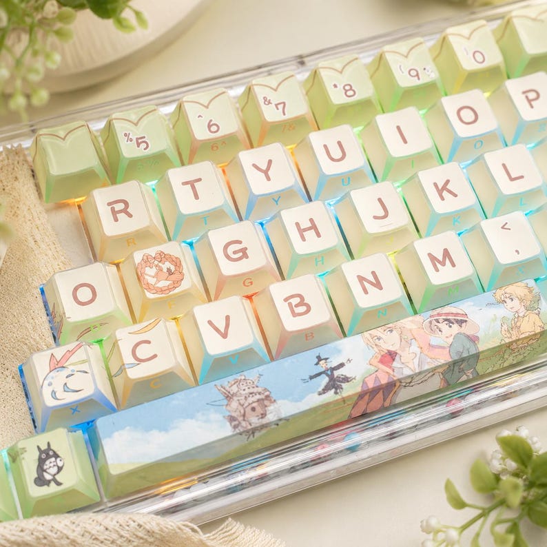 May include: A mechanical keyboard featuring light green and white keycaps with brown lettering. The spacebar displays a colorful illustration of a castle, a scarecrow, and two characters. Additional keycaps feature small character graphics.