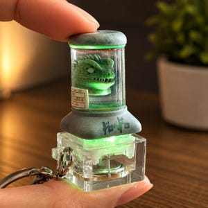 Peut inclure: Un porte-clés en acrylique transparent pour clavier mécanique avec une chambre cylindrique contenant une petite tête de créature reptilienne verte. La base brille d'une lumière LED verte vive et le côté du cylindre affiche une petite étiquette blanche.