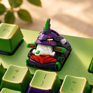 May include: A mechanical keyboard keycap featuring a stylized purple, green, and white robot head design with red accents. The keycap is mounted on a green keyboard base with other green keys visible, including one marked with the text F11.