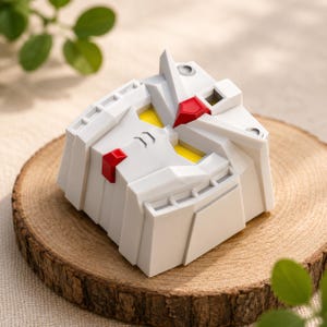 May include: A white mechanical robot head keycap featuring a geometric design with yellow eye accents, a red forehead V-fin center piece, and a small red chin detail. The object has a structured, angular appearance with recessed vents and panel lines.