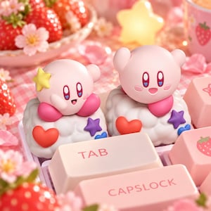 May include: Two pink Kirby character keycaps featuring a cloud-like base decorated with red hearts and purple stars. One figure has a yellow star on its head. The keycaps are positioned among light pink keyboard keys labeled TAB and CAPSLOCK.