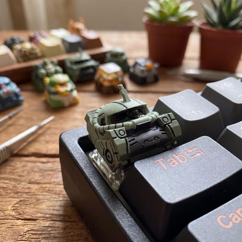 May include: A custom artisan mechanical keyboard keycap shaped like a sage green robotic head with a black visor and antenna, mounted on a black keyboard next to a Tab key. Several other detailed miniature character keycaps are visible in the background.