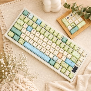 May include: A mechanical keyboard featuring a white frame and a custom keycap set in shades of sage green, light blue, and cream. The keys display QWERTY layout letters in red, along with various small line-art illustrations of woodland creatures.
